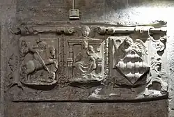 a bas relief carved stone with three sections including a man on a horse attacking a dragon, a woman with child seated, and a guardian angel figure behind a diamond-shaped shield