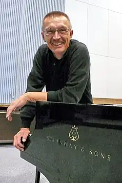 Emil Viklický leaning on a Steinway piano and grinning at the camera