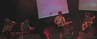 Three guitarists, a keyboard player, and a drummer are performing a song live on a stage lit by blue background sheets.