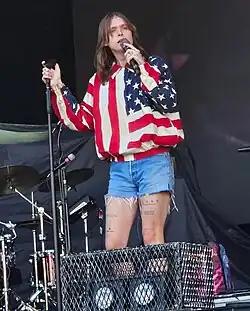 Cain singing into a microphone wearing an American flag-themed shirt and jean shorts