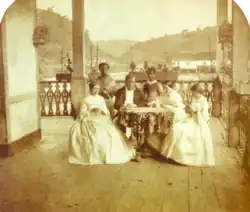 a man and three women seated around a table with two servants standing in the background