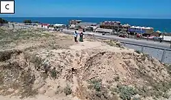 Two people stood on top of a yellow rocky mound. There is a road in the background with buildings on one side and the sea beyond that.