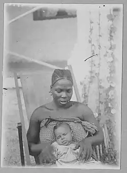Black-and-white photograph of a black woman sitting on a lounge chair holding a baby on her lap. The woman looks at the baby. She has beaded hair and facial scars.She wears a print cloth wrapped around her body. The baby has its eyes closed and wears a striped gown. The baby's hair is straighter than hers.