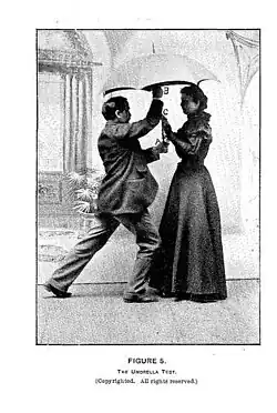A person stands under and forcefully holds an umbrella while Hurst stands under it and places her hands on it.