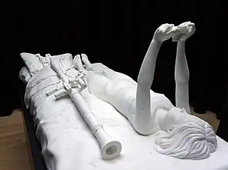 Foto of life size statuario marble sculpture with a black background. The sculpture is of a bed, with a naked young woman laying down, taking a selfie with a photo camera. Beside her on the bed lay large military weapons