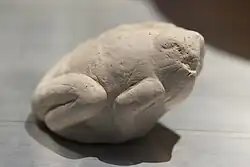 A light-grey stone shaped to resemble a crouchinh frog. The head is eroded, but the limbs are still visible tucked under the body.