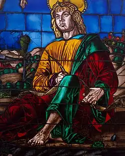 Detail of a stained glass window depicting a saint dressed in yellow, green and red robes, holding a quill.
