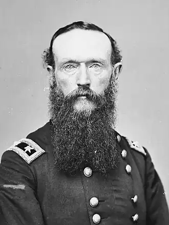 Black and white photo shows a heavily-bearded man staring directly into the camera. He wears a dark, double-breasted military uniform with general's stars on the shoulder tabs.