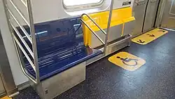 The interior of an R211 car, showing a stationary blue bench next to three foldable yellow seats. The yellow seats are shown here in the folded-up position.