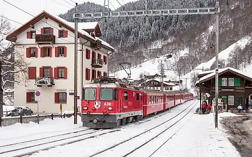 Red train arrives on snow-covered tracks at a two-story building with gabled roof