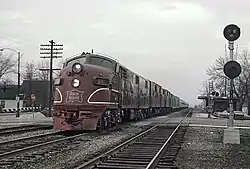 A red electric locomotive approaches the viewer from the left lane in a right-hand-drive context, opposite of the viewer. A telephone pole, stop sign, and railroad crossing can be seen in the distance.