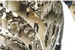 The same rotunda with the balconies covered in mirror-polished stainless steel. There are people walking along galleries, and a burly man with large beard, wearing, a black t-shirt, leans on the railing.