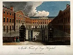 A view into a courtyard surrounded by a grand red brick building on three sides. Elaborate black railings cordon off the square on the fourth side, and patients are dotted about using crutches and being carried on stretchers.