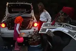 A parked white sedan is seen decorated with pirate-related Halloween decorations as a child in an orange wig takes candy from the trunk of the vehicle; a skeleton pirate statue holds the sign with the words "Walk the Plank FOR CANDY".