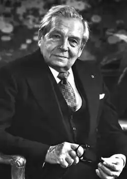 A black and white photo of jeweler Harry Winston sitting in a chair, looking into the camera and holding his glasses in his hands