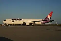 A white twin-engine plane painted with the word "HAWAIIAN" in the front above the windows, a black, silver, and white logo of a pirate's head on the forward bottom of the fuselage, and a woman in different purple hues on the tail taxis at an airport