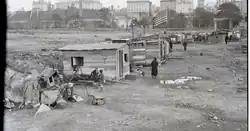 Hooverville with a few people around the wooden shanties, buildings are in the distance