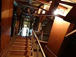 A wooden stairway surrounded by a complicated wooden structural system, rising under a curved wooden ceiling to a distant lit area, illuminated by a bare light bulb