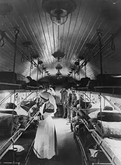 black and white image of a long train carriage, with three bunk levels of beds along the length of each side of the carriage. Some of the beds have patients. In the centre of the image, in the foreground, there is a woman in a nurse uniform, with long skirts and an apron, and a nursing habit on her head. She is faced to the left, tending to a patient in the middle bunk, in the background at the end of the carriage are soldiers are two soldiers standing on the right side facing to the left, one of the soldiers has his hand raised in a salute.