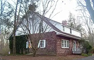 A house built of red stone house with a curved, sloping roof and white wooden top, seen from the side. It has brick chimneys at either end and some ivy on the visible side.