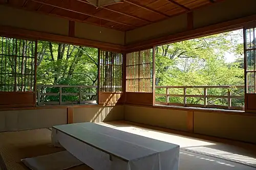 room with sliding screens at about 50&nbsp;cm height, looking out on the upper reaches of sunlit trees. The screens have a low wooden panel, and a high lattice which appears to be open but is actually glass-filled.