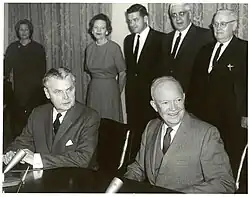 Diefenbaker and a smiling bald man in a suit sit at a table. Two women and two men stand behind them.