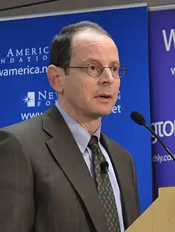 Rauch wearing a brownish grey plaid suit and round-rimmed eyeglasses, standing at a podium