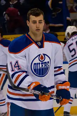 An ice hockey player in an orange, white, and blue Edmonton Oilers jersey standing up while holding his stick
