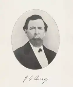 Black and white headshot of Joseph Avery. He's wearing a suit coat over a white shirt with small black tie (kind of like a bow tie). He has brown hair and a large chin-beard.