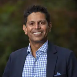 Karthik Krishnan wearing a white and light blue checkered shirt and dark blue blazer, with a light blue paisley pocket square, grinning directly at camera