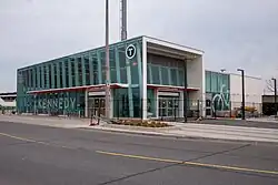 A photo of a glass panelled light rail station with the name "Kennedy" in bold white lettering on the side. The structure is mostly completed, but signage over the entrances has not yet been added.