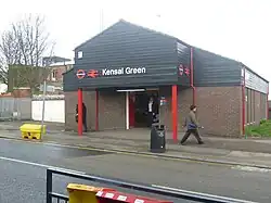 A brown-bricked building with a black sign reading "Kensal Green" in white letters and some people walking in front all under a white sky