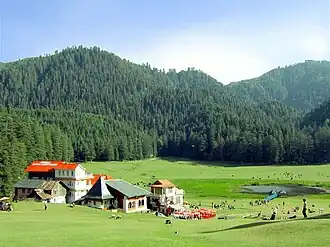 Daytime sunlit view of a verdant bowl-shaped meadow with a small pond in a valley rimmed by several prominent hills covered in evergreen forest. At centre-left and looking out on the meadow, a small cluster of tourist lodges with pitched roofs, one a bright red. Around them and the pond are small, gaily coloured gazebos and shade-giving umbrellas. Several dozen tourists and sheep mill about the pond and in the meadow.