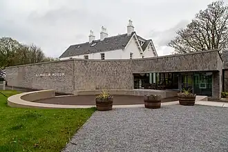 Exterior of Kilmartin Museum, showing its modern, curved greyish-brown brick facade with 'KILMARTIN MUSEUM' in light letters. A traditional white building with a dark slate roof and chimneys rises behind. A gravel path and barrel planters are in the foreground, under an overcast sky.
