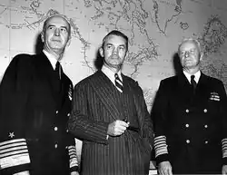 Black and white photograph of three men in front of a wall map. The one at the centre is in a striped suit and tie holding a pipe, both the men to his right and left are in a naval uniform jacket