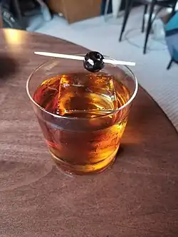 A beverage in a short glass with large ice cube and swizzle stick with cherry