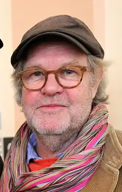 close-up of Kjell-Åke Andersson wearing a brown corduroy jacket, a colourful scarf, and a newsboy cap, looking directly at camera with a slight smile