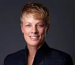 A professional portrait photograph of a white woman in a dark zippered top; she is smiling while facing and looking into the camera.
