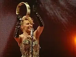 An image of Kylie Minogue, with one of her raising hands holding a tambourine. The microphone in a microphone stand is seen infront of Minogue.