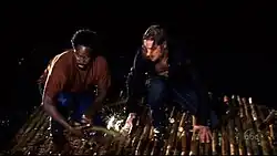 A black man and a white man, both wearing wet clothes, sit on a raft made of bamboo at night.