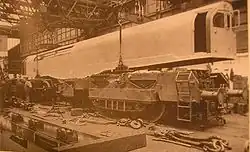 A picture of a locomotive under construction. The cuboid box containing cabs and boiler is being lowered onto the two bogies by a crane inside a locomotive works. Various components are scattered in the foreground.