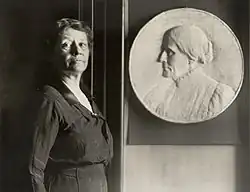 A woman emerging from shadow stands next to a large round bas-relief sculpture of Susan B. Anthony's head.