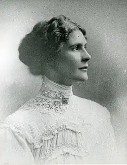 A sepia-toned portrait of a woman with wavy hair styled in an updo, wearing a high-collared lace blouse, gazing to the right with a serene expression.