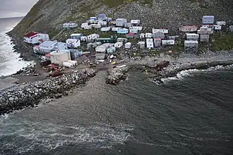 A village of 40–50 mostly small buildings built on a steep, rocky coastline; some structures higher on the hill are built on stilts