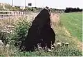 Brown triangular stone at edge of grassy field near fence and bank of motorway