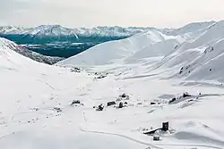 "A top-down view of the site, showing approximately 10 wooden buildings in the snow. Groomed cross-country ski trails weave through and around the buildings."