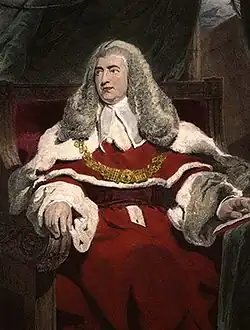 A middle age man sits in judicial robes, with a judge's wig on his head