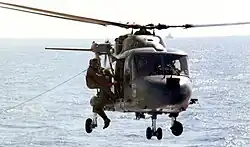 Two men in flight suits grapple on the side of a dark blue helicopter hovering over water.