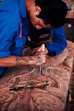 A craver in a blue jacket carving a traditional Māori wood carving