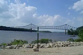 A steel cantilever-truss bridge arches gracefully over a tree-lined river.